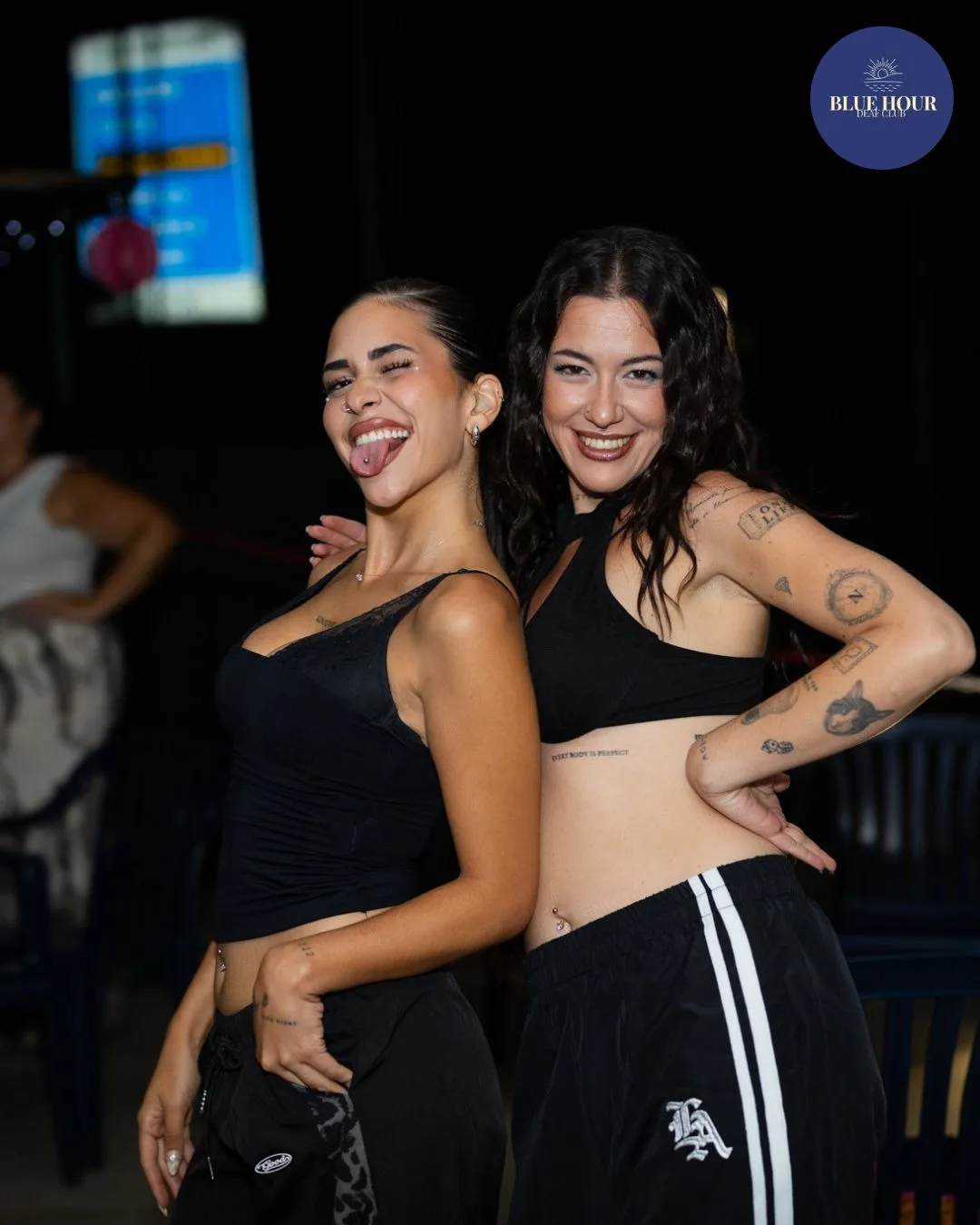 Due amiche in nero ridono abbracciate, una fa la linguaccia